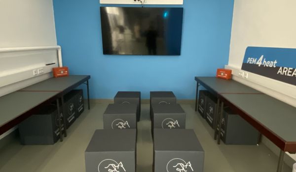 An einer blauen Wand hängt ein Fernseher. Davor stehen Tische und Hocker.