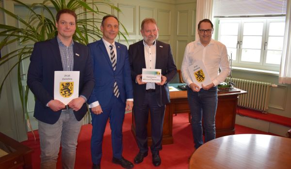 Vier Männer stehen in einer Reihe nebeneinander. Zwei von ihnen halten eine Urkunde. Ein Mann hat ein Schild zur MINT Kooperation.