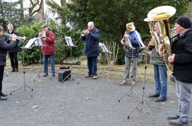 Personen mit verschiedenen Blechblasinstrumenten stehen draußen im Halbkreis und musizieren.