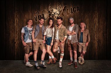 Eine Volksmusikgruppe in Lederhosen posiert vor einer Holzwand mit der Aufschrift: Die VolXXLiga.