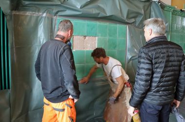 Drei Männer stehen vor einer grünen Fliesenwand und begutachten diese.