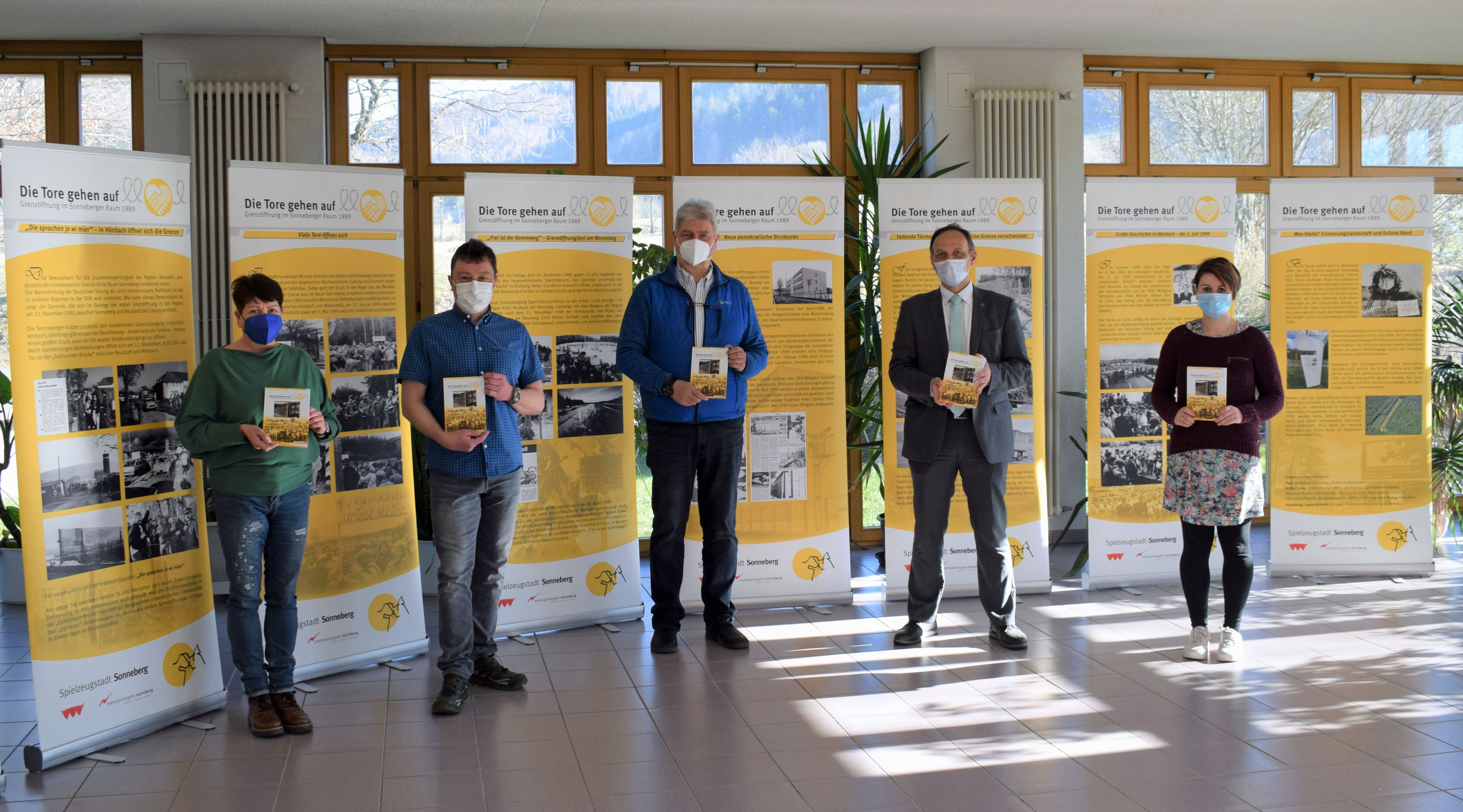 Fünf Personen stehen vor verschiedenen Bannern. Sie halten ein Buch in der Hand.