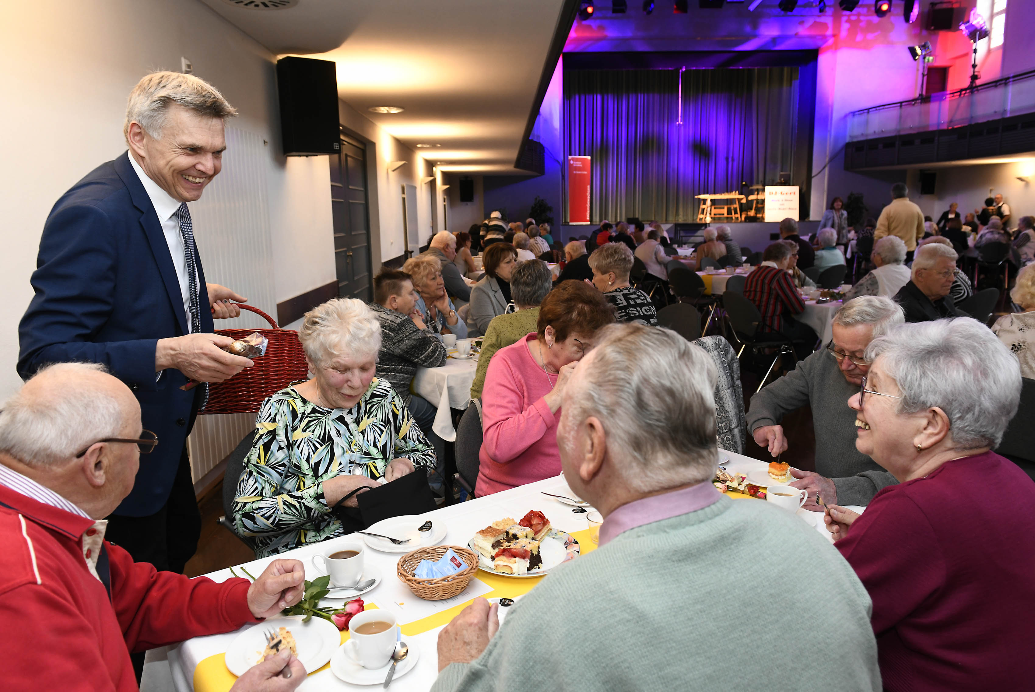 Menschen sitzen an einer gedeckten Kaffeetafel. Ein Mann teilt Geschenke aus.