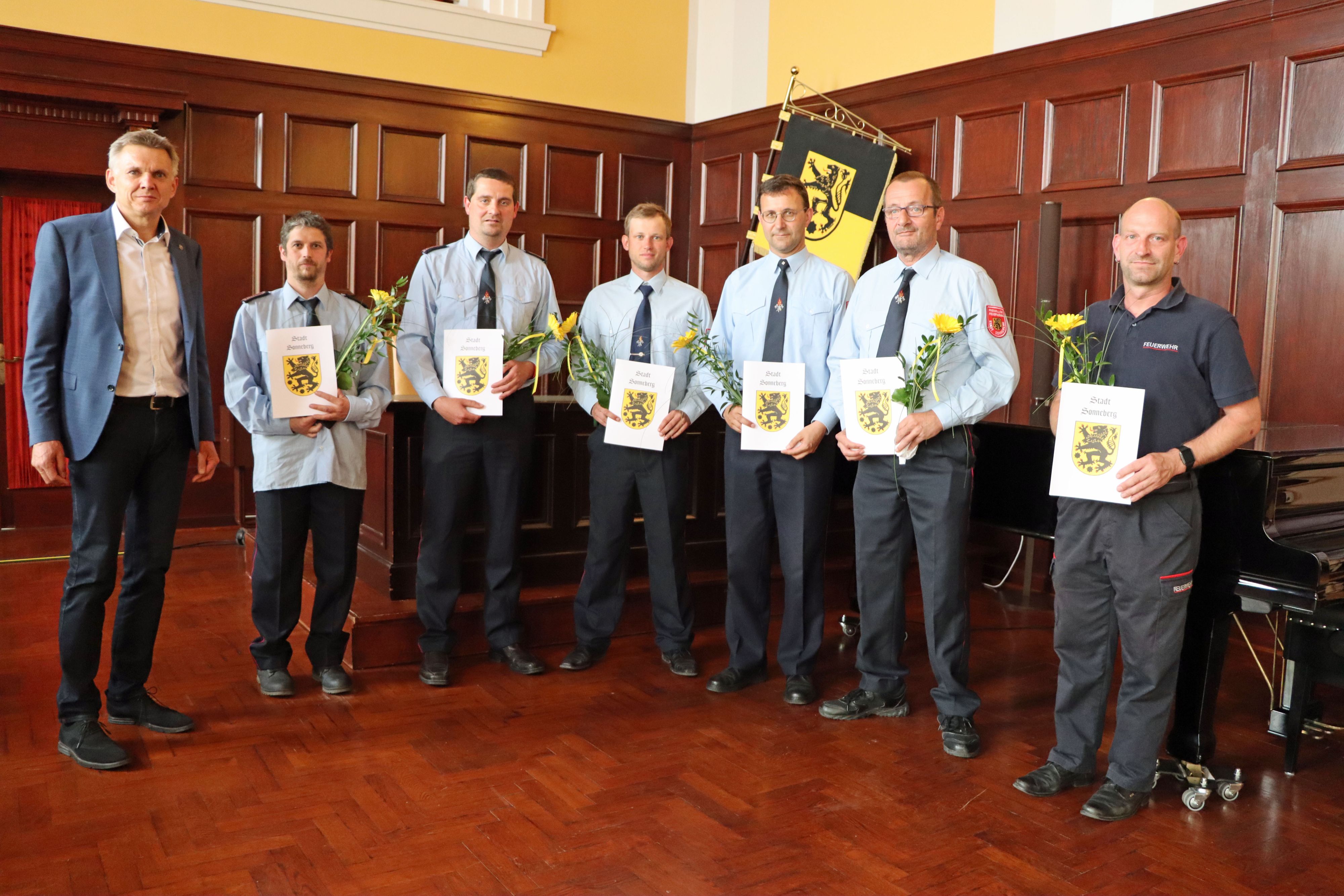 Männer in Feuerwehrunifrom stehen in einer Reihe nebeneinander. Sie halten eine Urkunde und eine Blume in der Hand. Links daneben steht ein Mann im Anzug. Es ist der Bürgermeister.