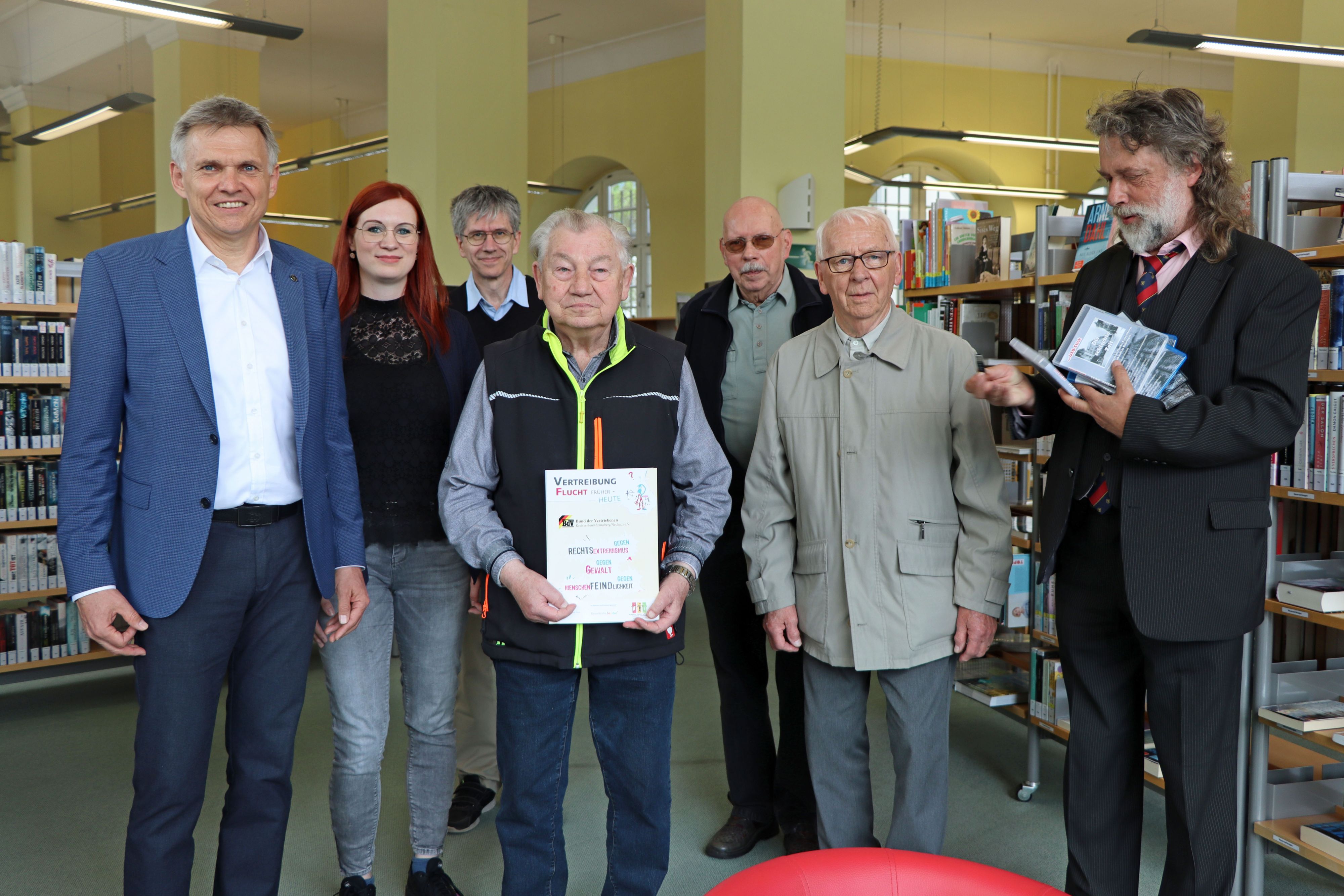 Mehrere Personen stehen nebeneinander. Ein Mann in der Mitte hält ein Poster mit der Aufschrift: Vertreibung, Flucht - früher bis heute. Ein anderer Mann hält DVD-Schachteln in der Hand.