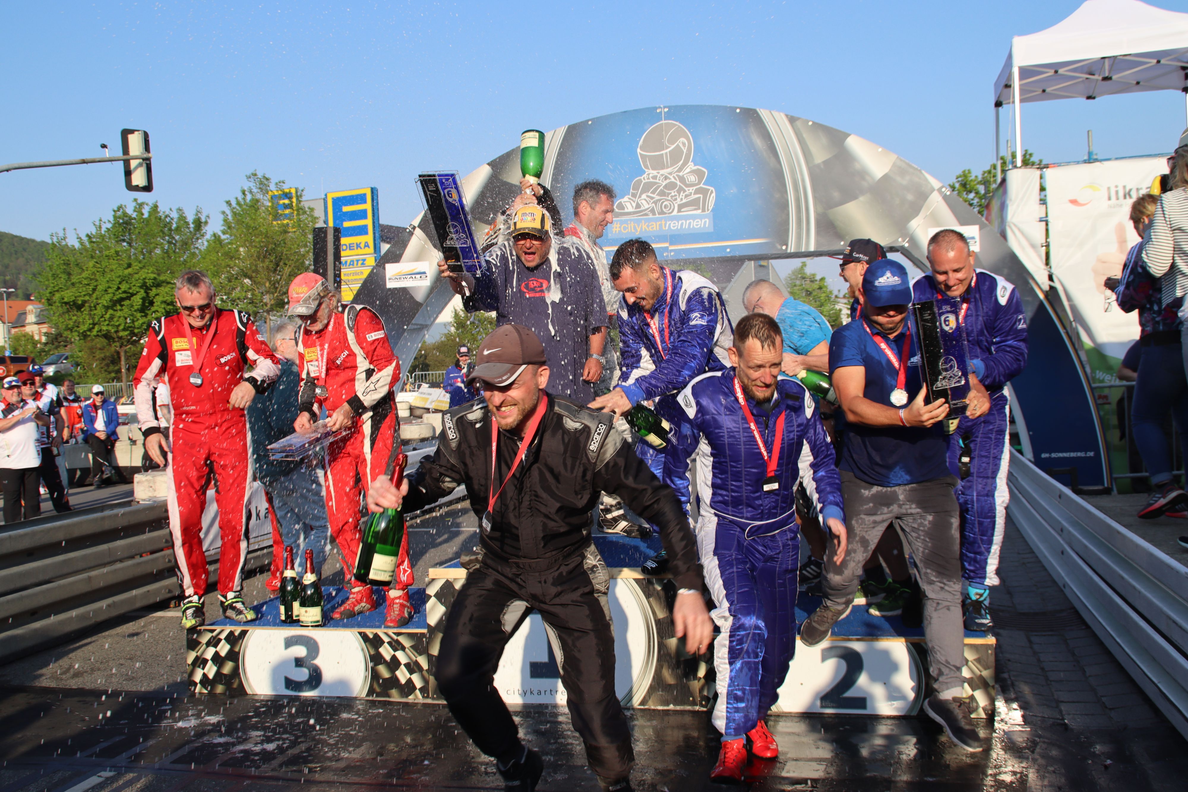 Mehrere Männer in Rennanzug stehen auf einem Siegerpodest. Sie tragen eine Medaille um den Hals. Ein Mann bekommt eine Flasche Sekt über den Kopf geschüttet.