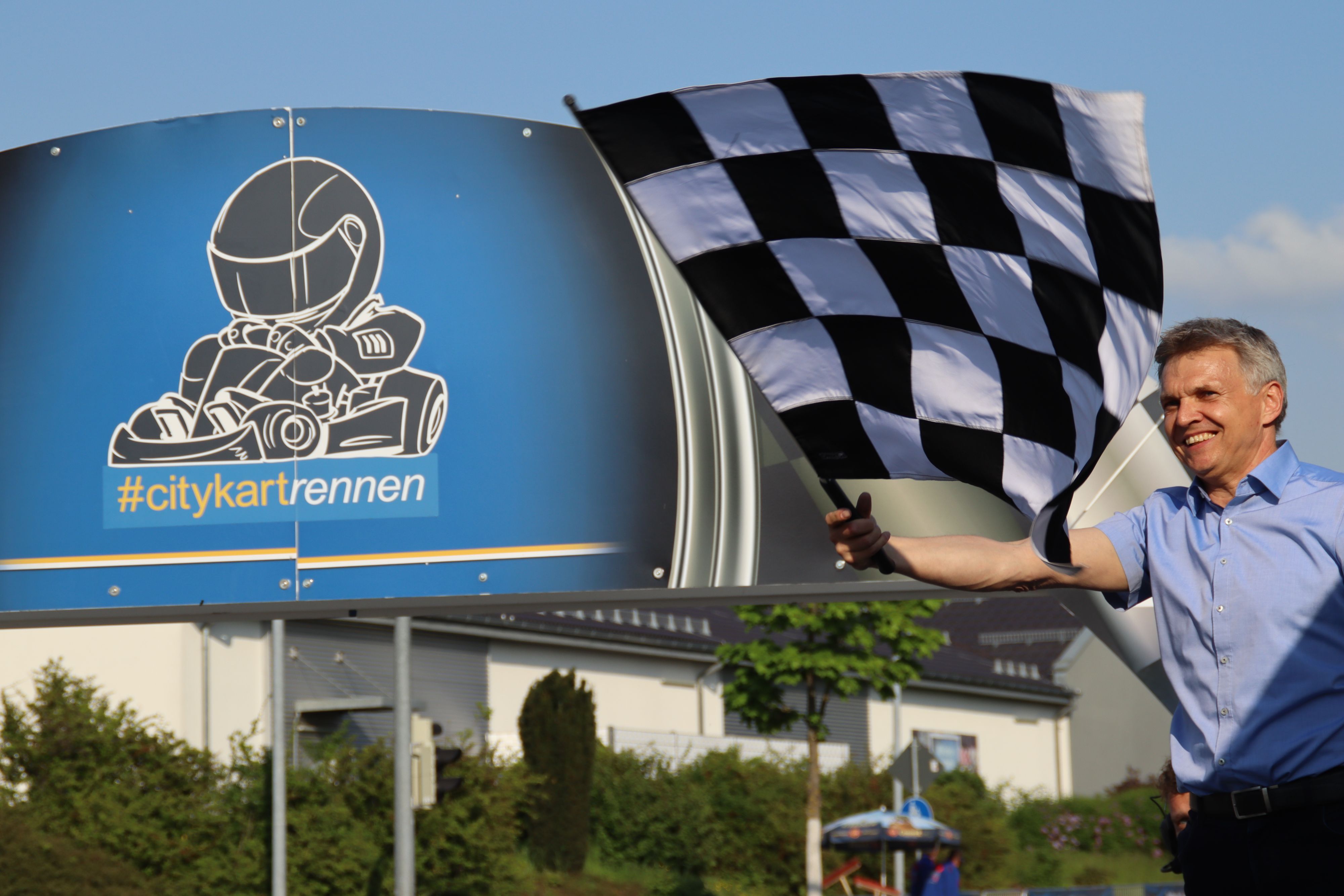 Ein Mann schwingt eine schwarz-weiß karierte Flagge. Es ist der Bürgermeister Dr. Heiko Voigt. Dahinter steht ein Schild mit der Aufschrift: #citykartrennen.