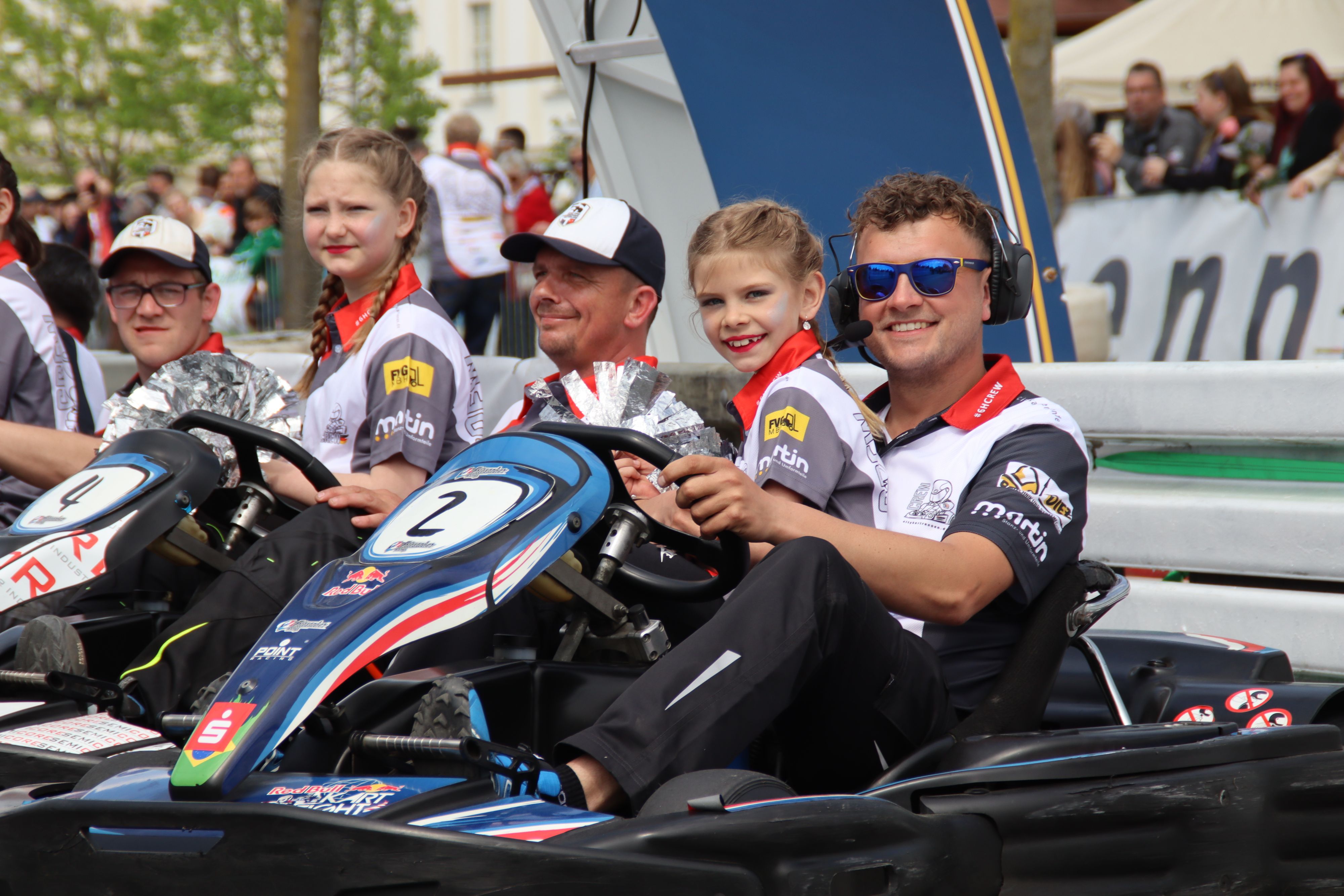 Männer sitzen mit einem Kind auf dem Schoß in Kartautos nebeneinander.