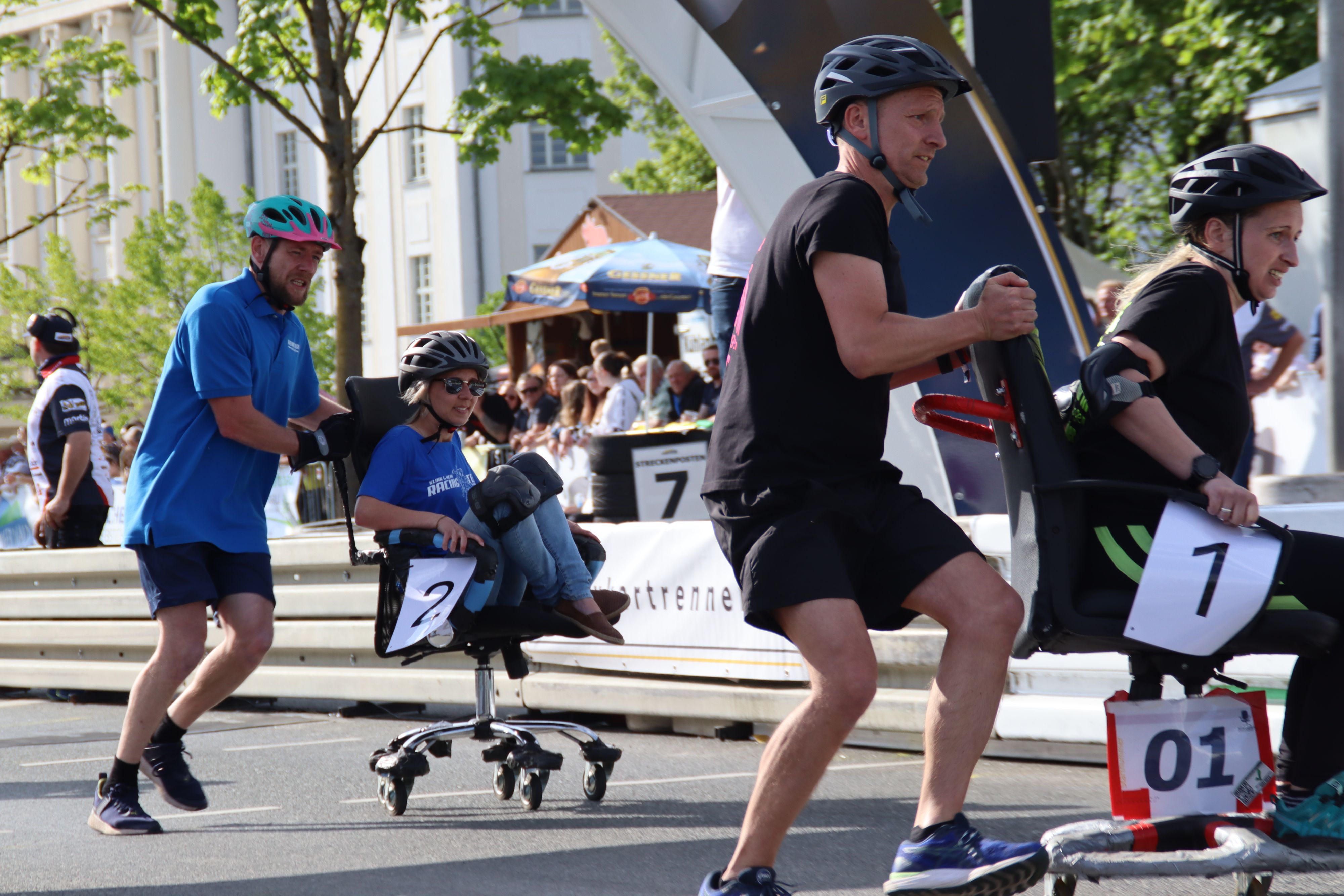 Zwei Frauen sitzen auf Bürostühlen und werden von zwei Männern über eine Rennstrecke geschoben. Im Hintergrund stehen Zuschauer.