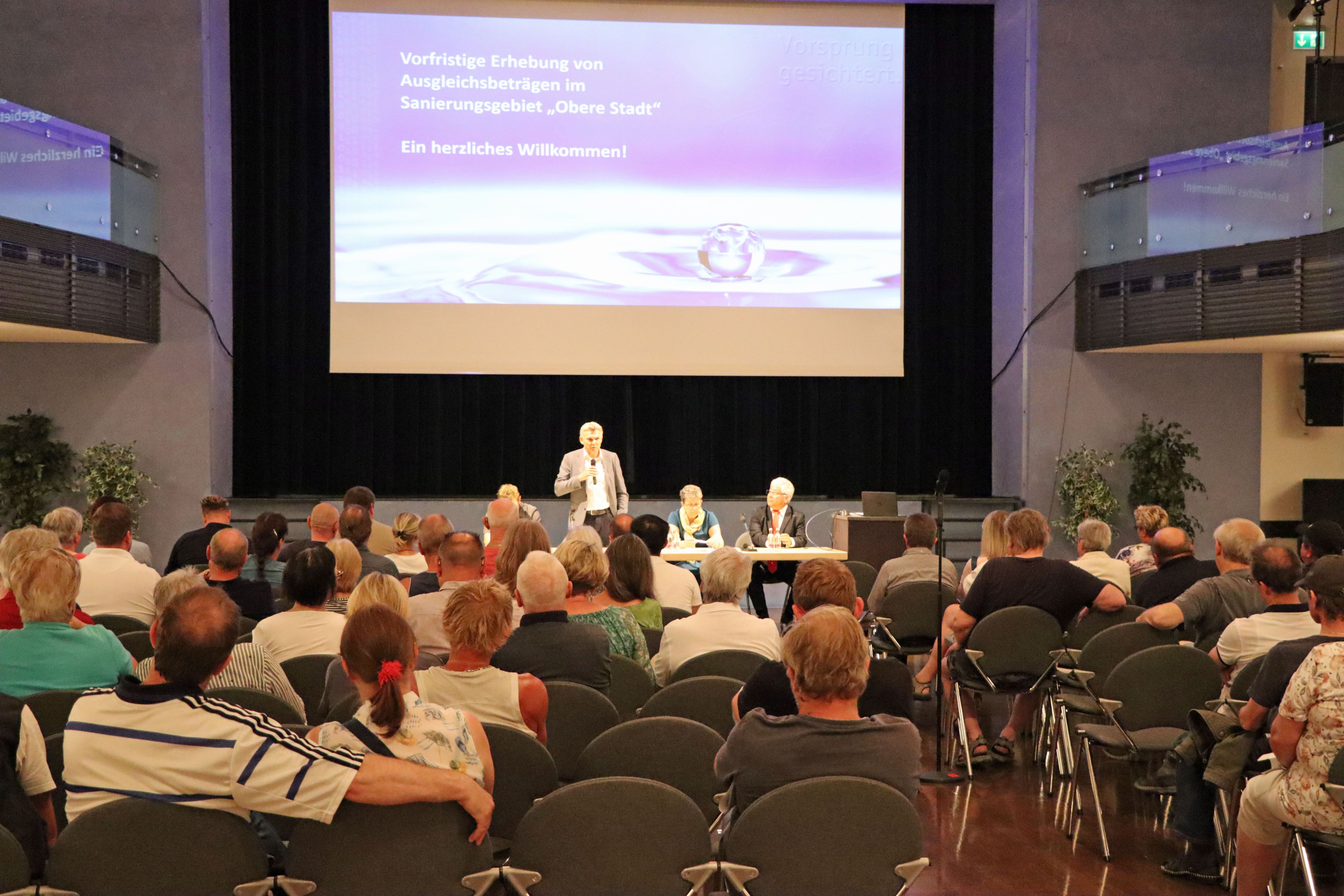 Menschen sitzen in Stuhlreihen vor einer großen Beamer-Leinwand. Vorne steht ein Mann. Es ist der Bürgermeister der Stadt Sonneberg.