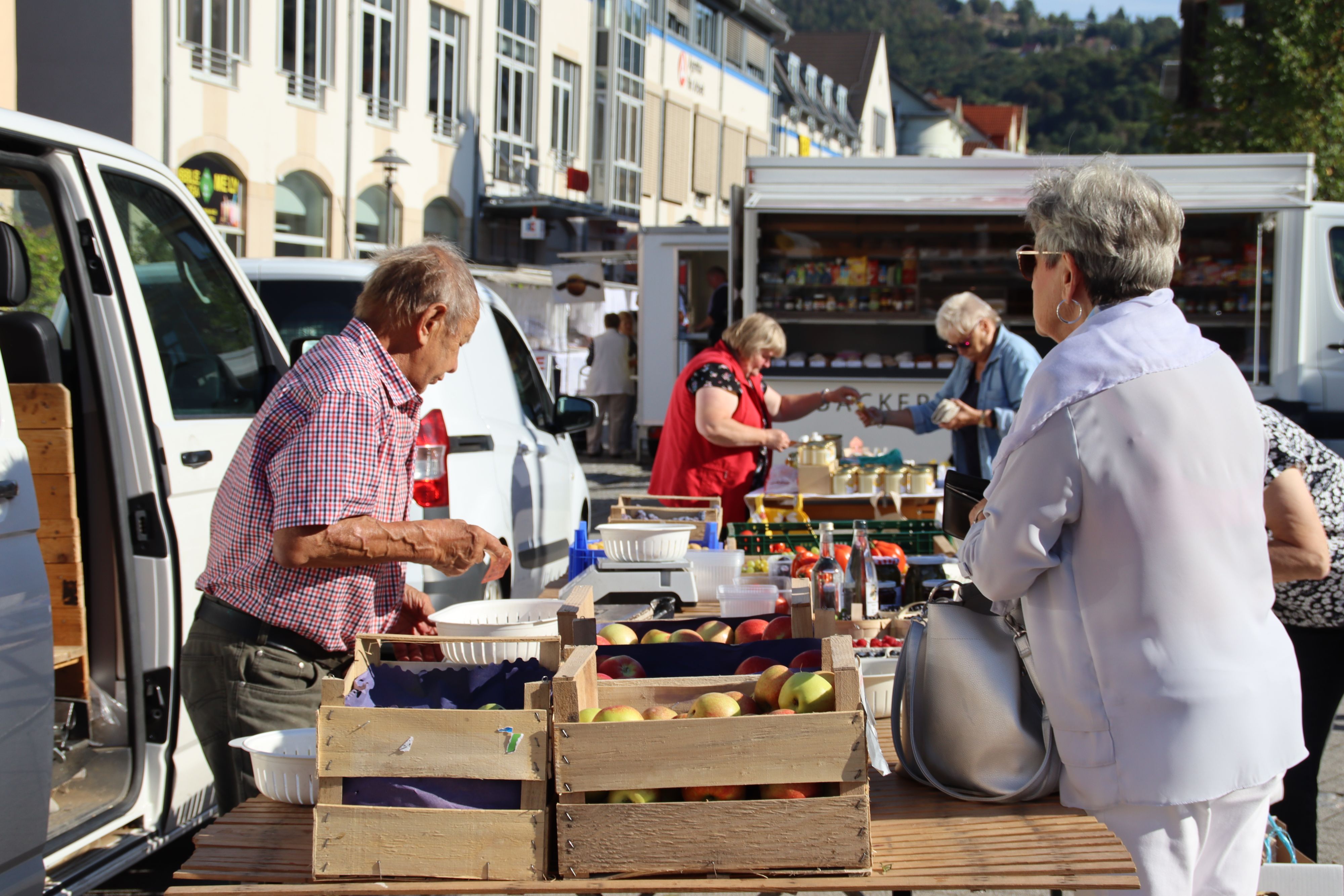 Auf einem Tisch stehen Holzkisten mit Obst und anderen frischen Lebensmitteln darin. Hinter dem Tisch steht ein Verkäufer. Vor dem Tisch steht eine andere Person mit einem Geldbeutel in der Hand.