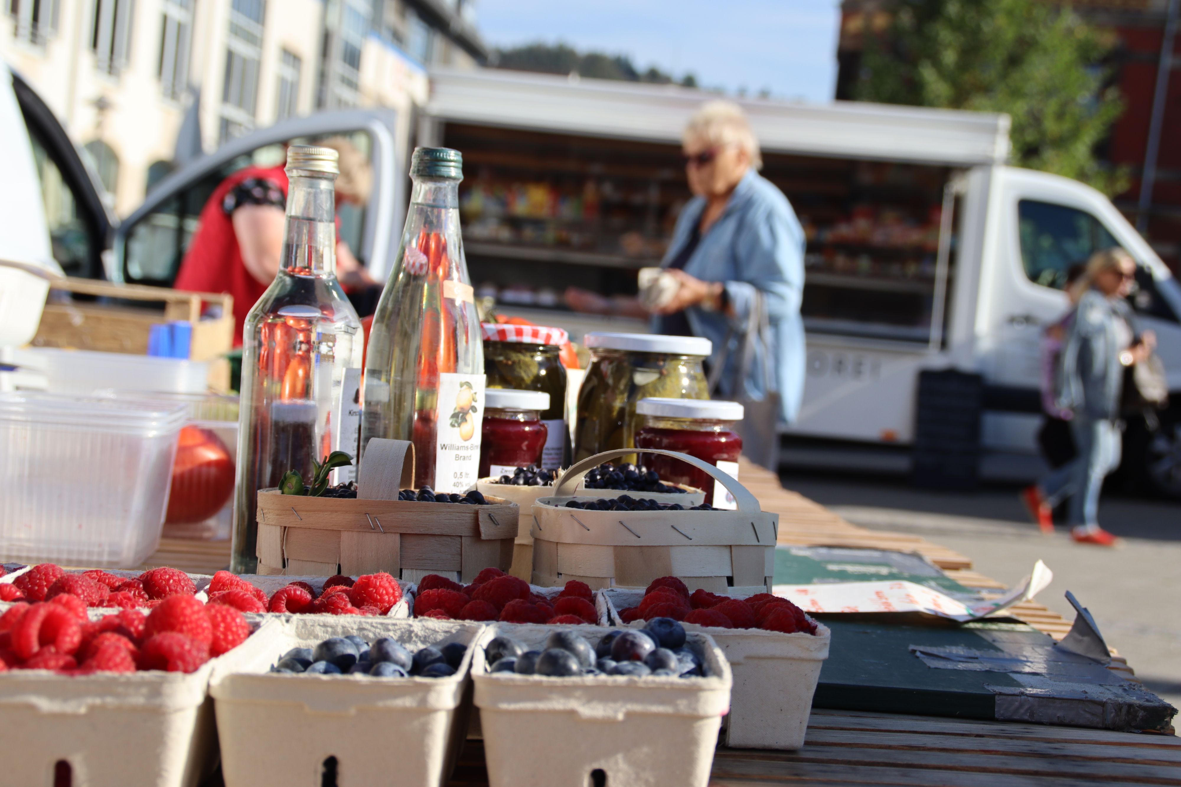 Auf einem Tisch stehen Kisten mit Obst und Selbstgemachtes. Im Hintergrund steht eine Frau mit einem Geldbeutel in der Hand.