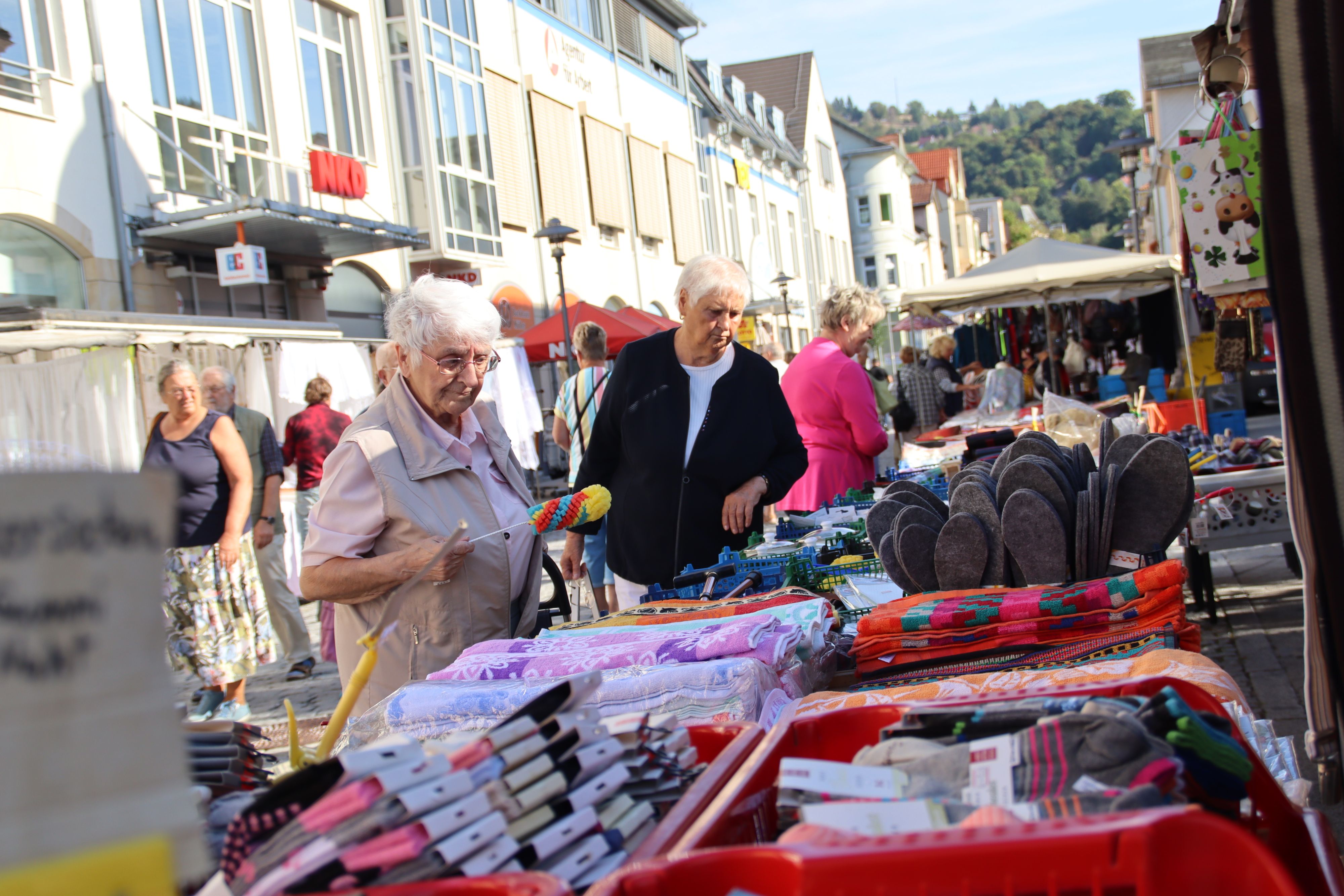 Auf einem Tisch stehen klappbare Kisten. In den Kisten sind Socken, Hausschuhen und Handtüchern. Vor dem Tisch stehen interessierte Kunden.