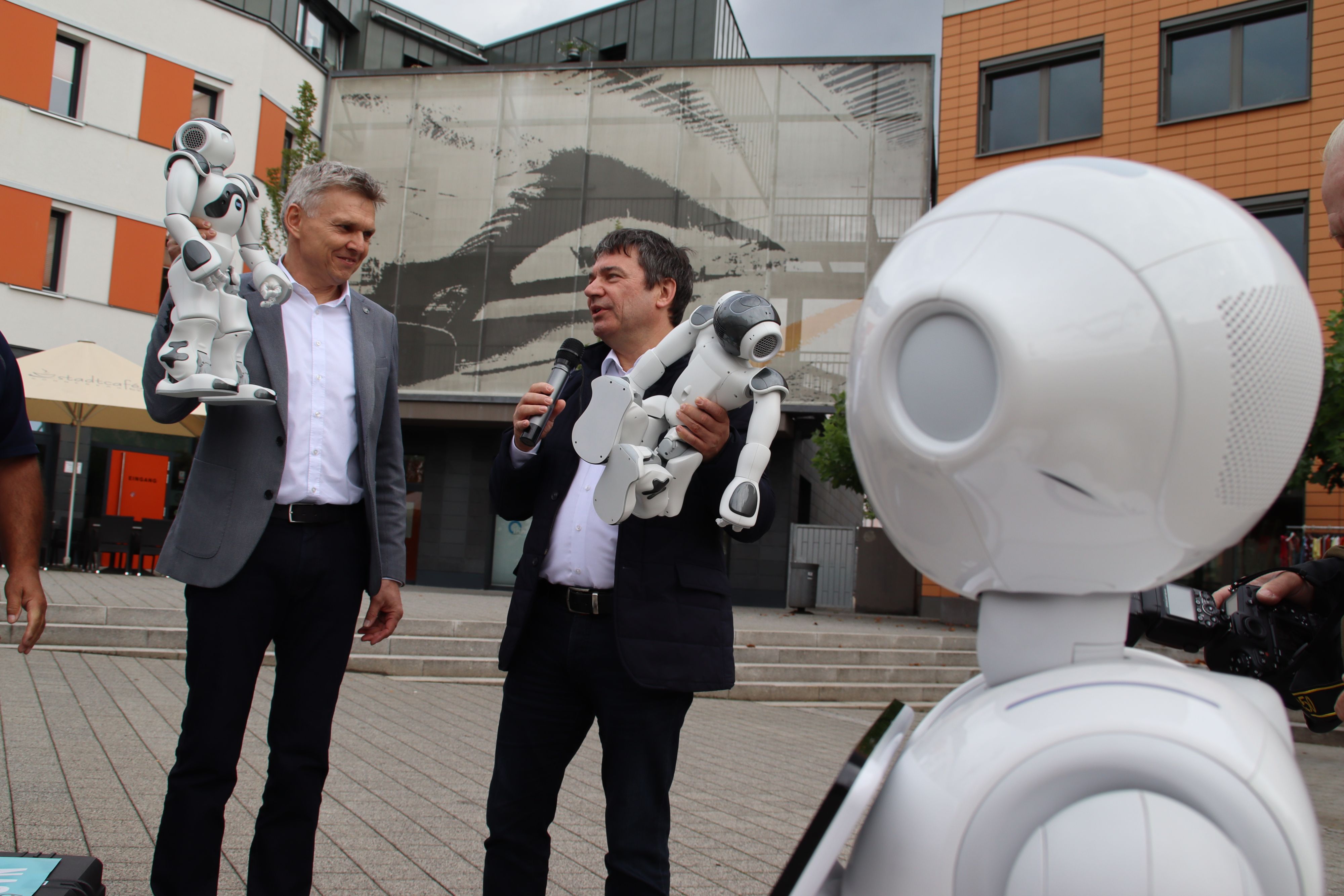Zwei Männer im Anzug stehen in der Innenstadt von Sonneberg und halten jeweils einen Roboter in den Händen. Im Vordergrund steht ein größerer Roboter.