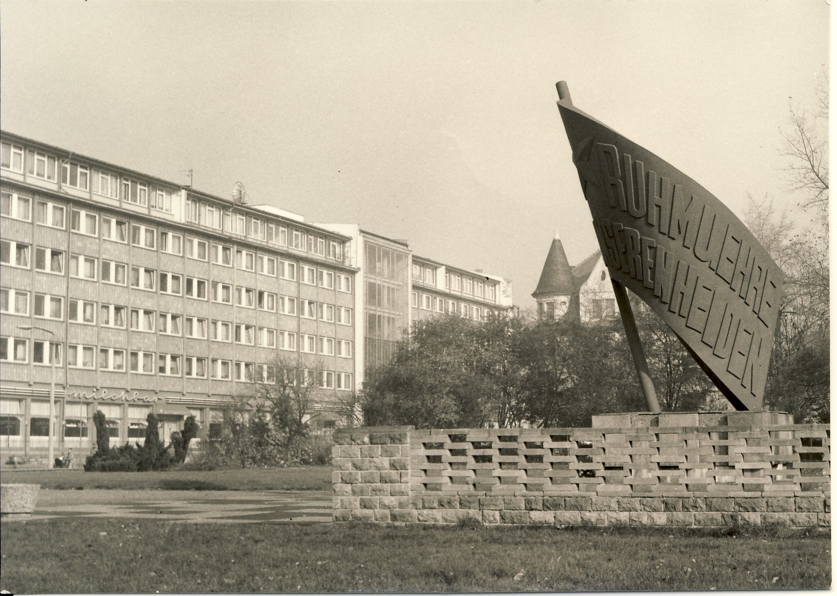 Schwarz-weiß: Ein Flaggendenkmal aus Stein mit der Aufschrift: Ruhm und Ehre unseren Helden. Im Hintergrund steht ein großes Gebäude.