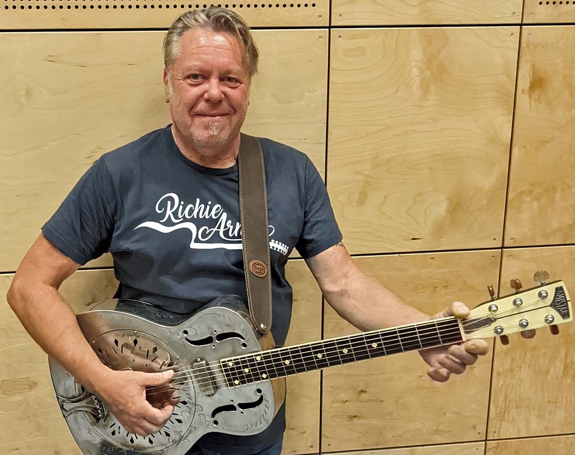 Ein Mann hält eine Gitarre aus Metall in der Hand. Hinter ihm ist eine Holzwand.