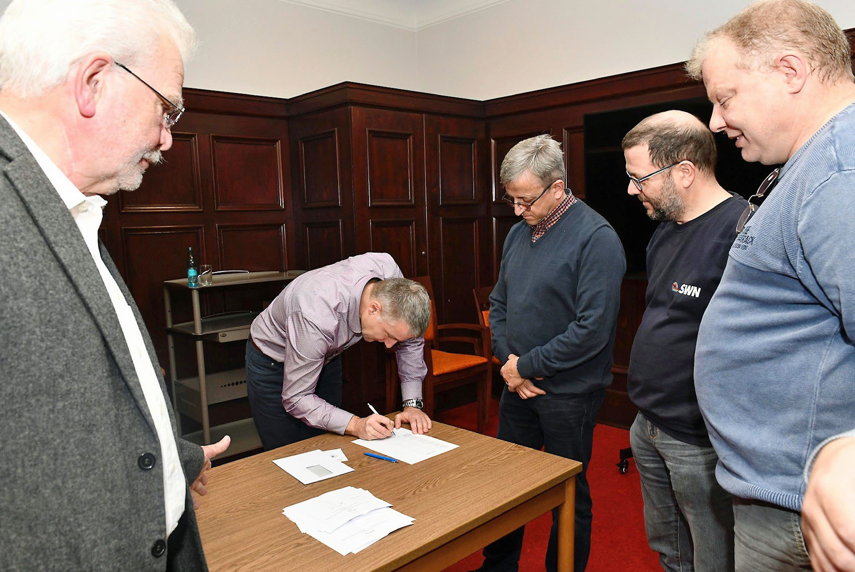 Fünf Männer stehen um einen Tisch herum. Einer der Männer beugt sich nach vorne und schreibt etwas auf ein Blatt Papier.