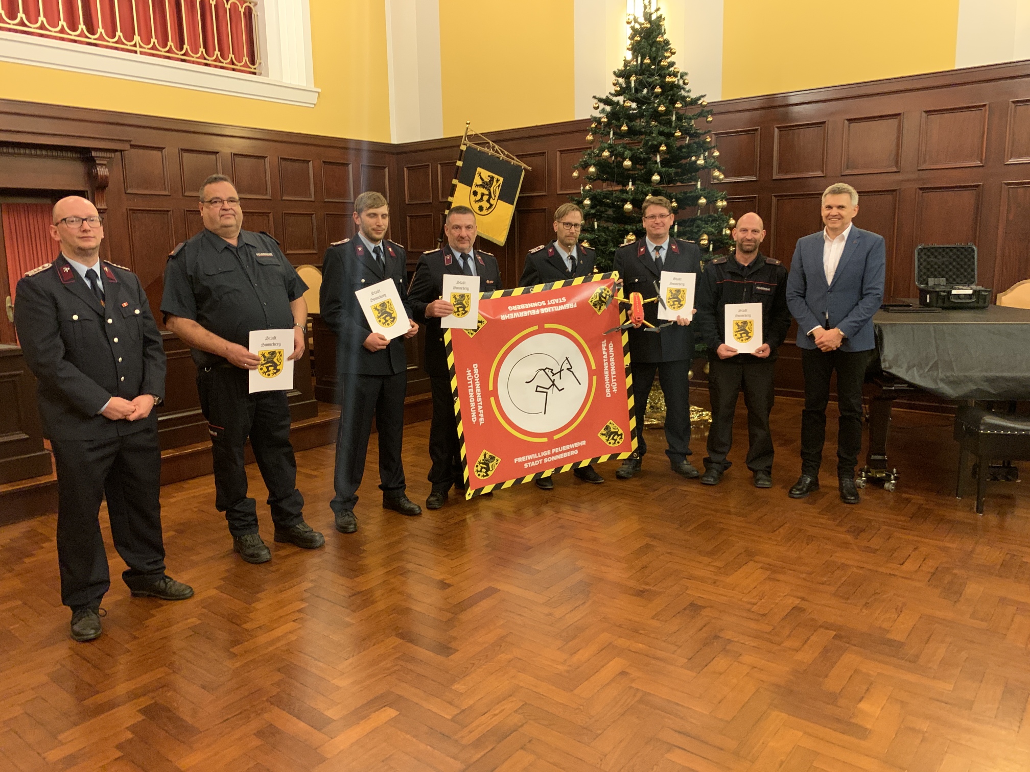 Sieben Männer in Feuerwehr-Uniform stehen in einer Reihe nebeneinander. Fünf von ihnen halten eine Urkunde. Rechts daneben steht der Bürgermeister Dr. Voigt.