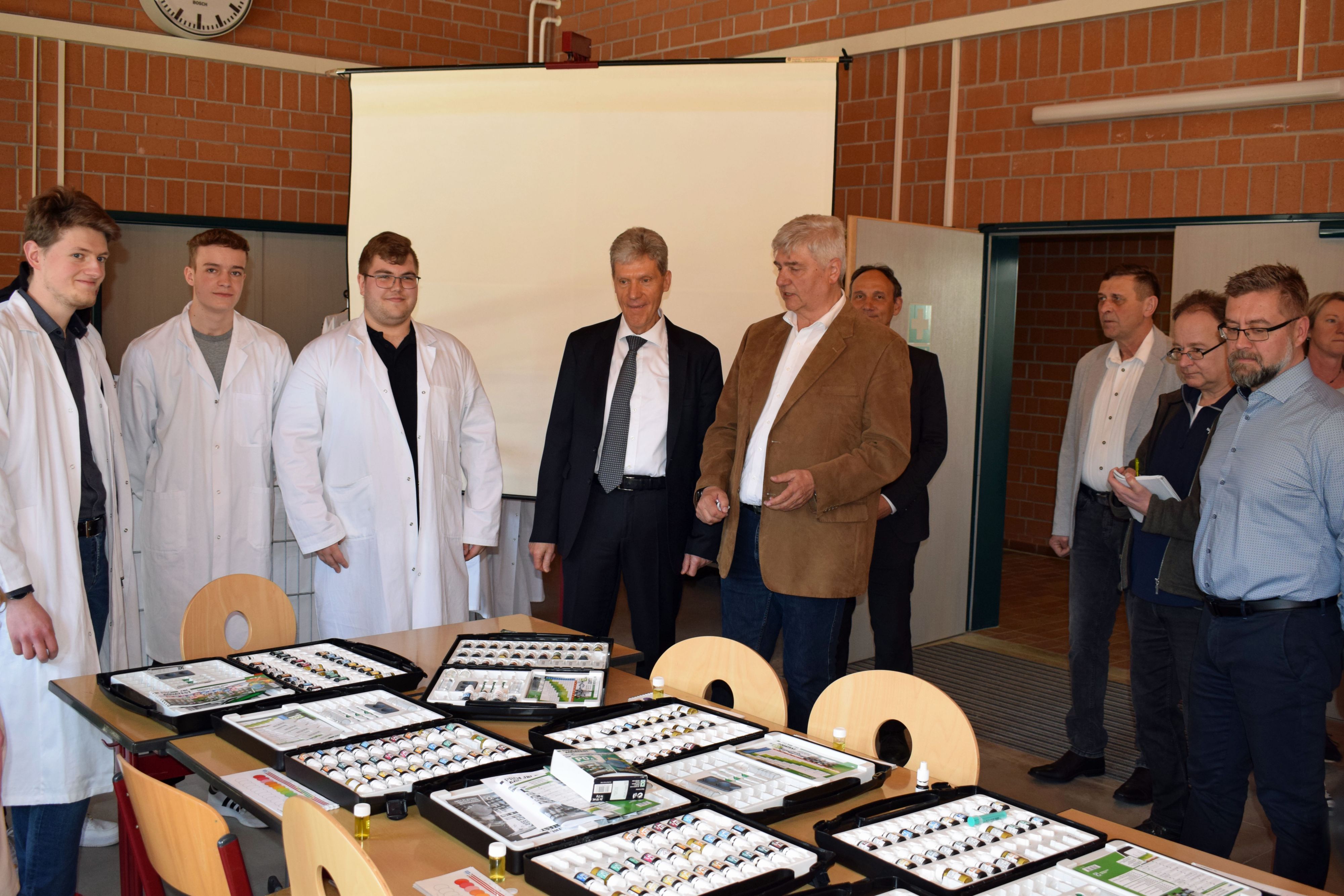 Auf einem Tische liegen Experimentierkoffer. Links stehen Jugendliche in weißen Kitteln. Rechts stehen Männer im Anzug.