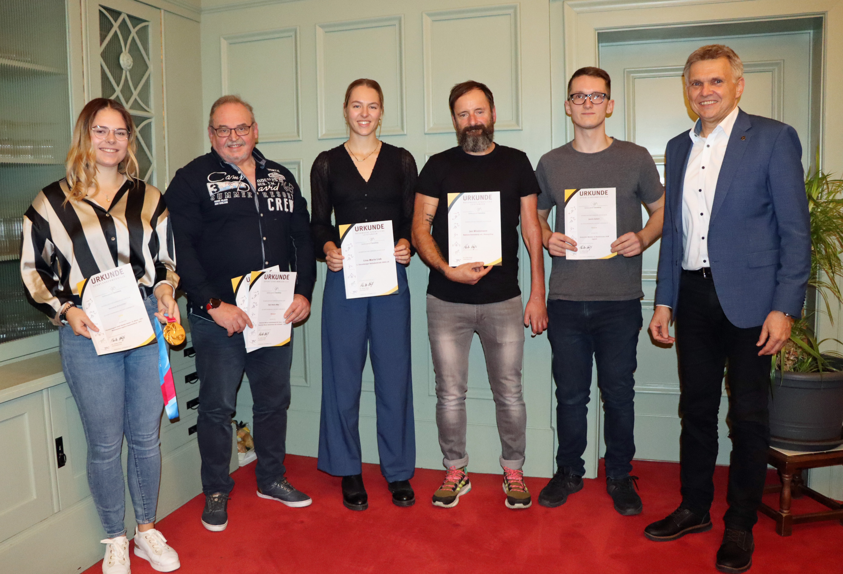 Fünf Personen stehen in einer Reihe nebeneinander. Sie halten eine Urkunde in den Händen. Rechts daneben steht der Bürgermeister.