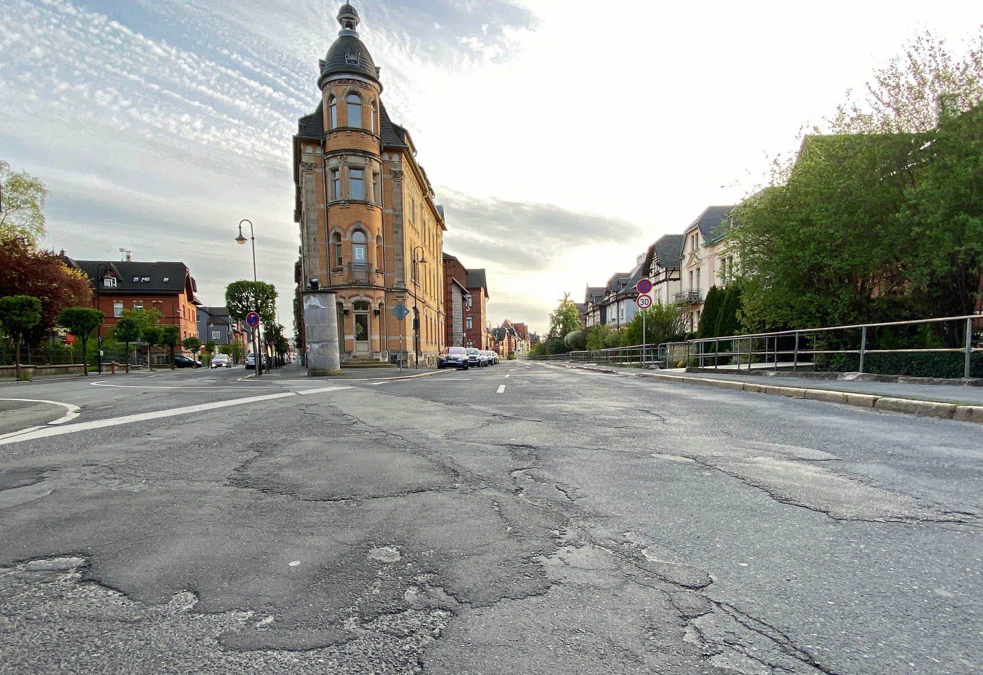 Es ist eine Straße zu sehen mit vielen Flicken, Rissen und Löchern. Im Hintergrund stehen Häuser.