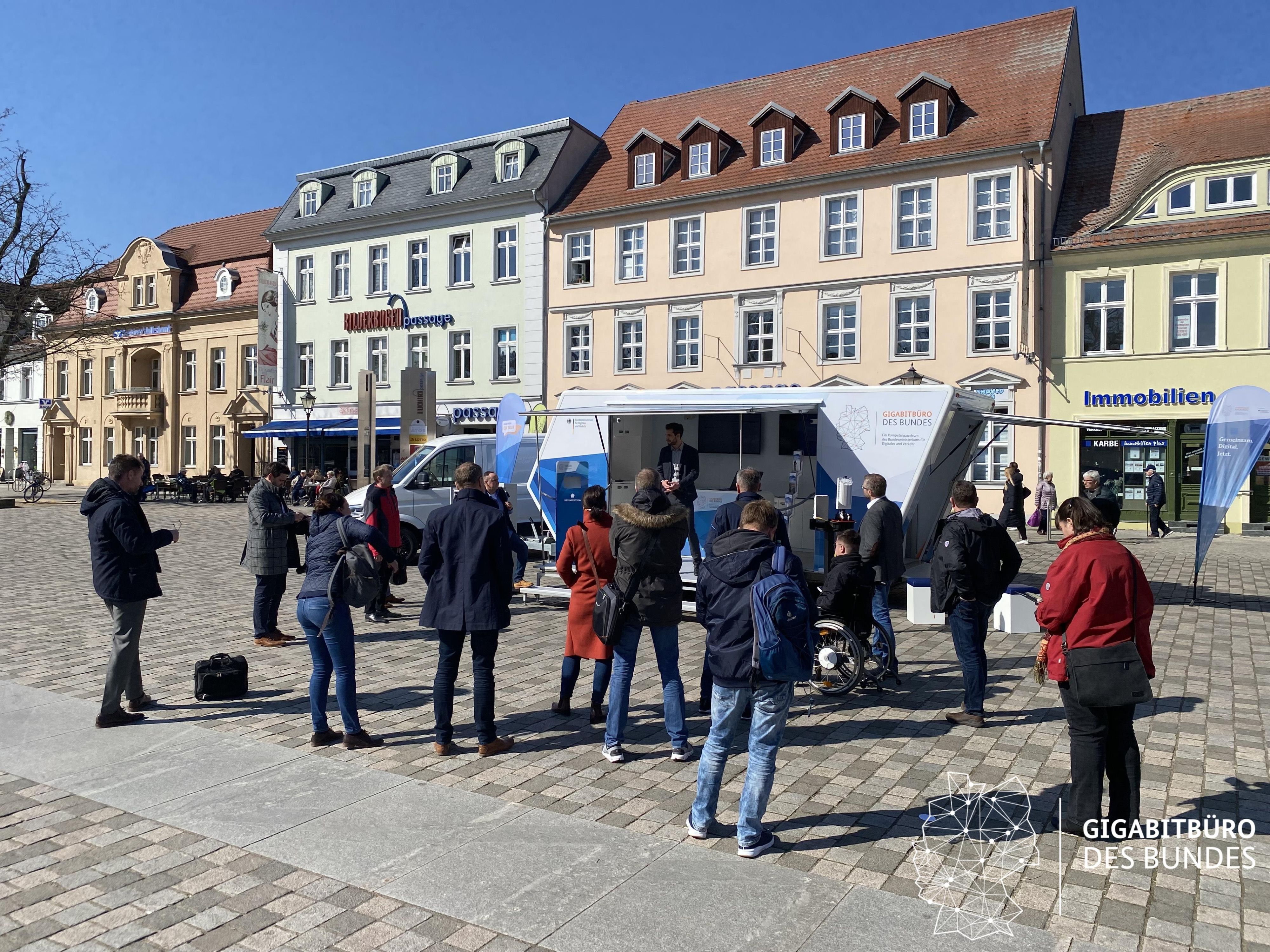 Eine Traube von Menschen steht auf einem Marktplatz vor einem geöffneten mobilen Wagen mit Informationen zum Thema Internet.