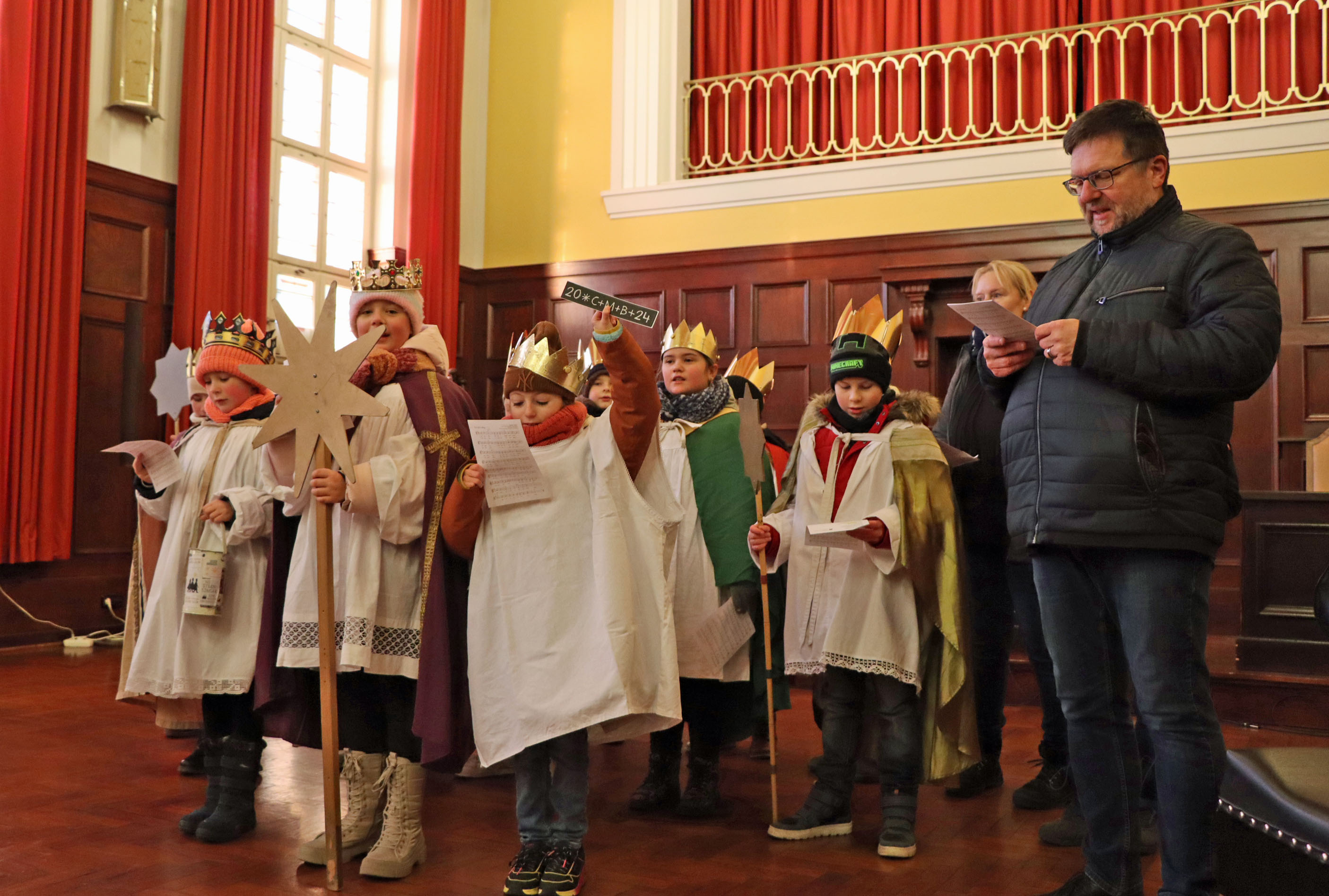 Kinder sind als Sternsinger gekleidet. Sie stehen in einem Saal und singen.