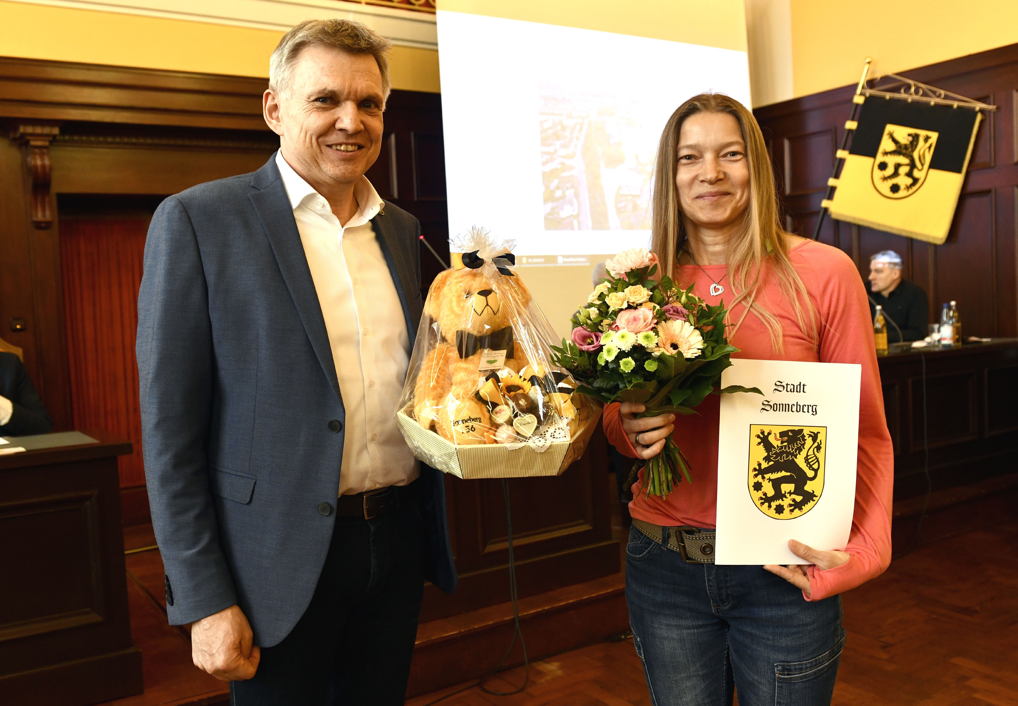 Eine Frau ält eine Urkunde und einen Blumenstrauß und bekommt vom Bürgermeister noch einen Teddy überreicht.