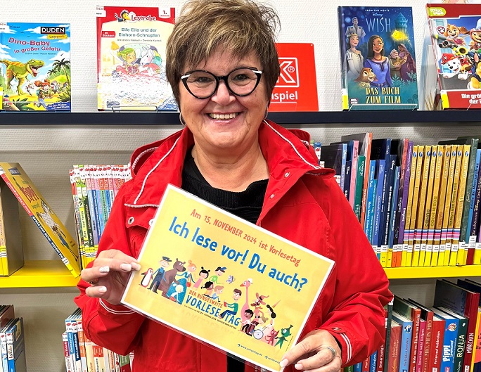 Eine Frau hält vor einem Bücherregal ein Schild in die Kamera. Darauf steht: Ich lese vor! Du auch?