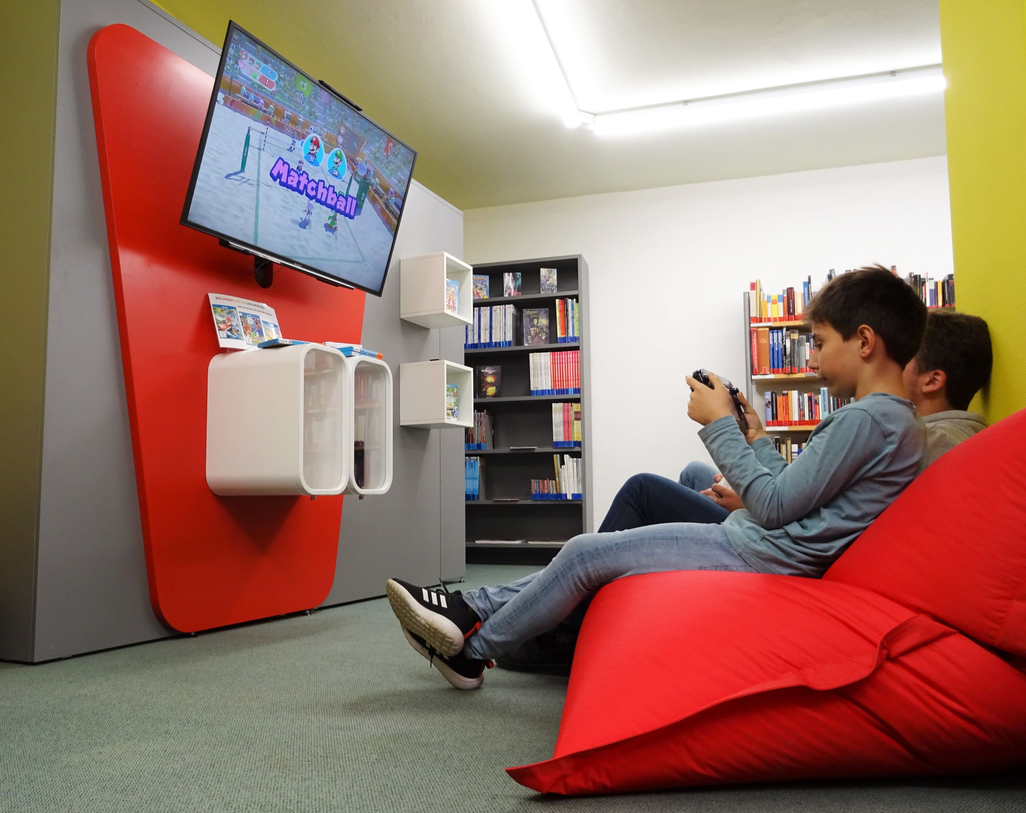 Zwei Jungs sitzen auf roten Sitzkissen vor Bücherregalen und schauen in einen Bildschirm.