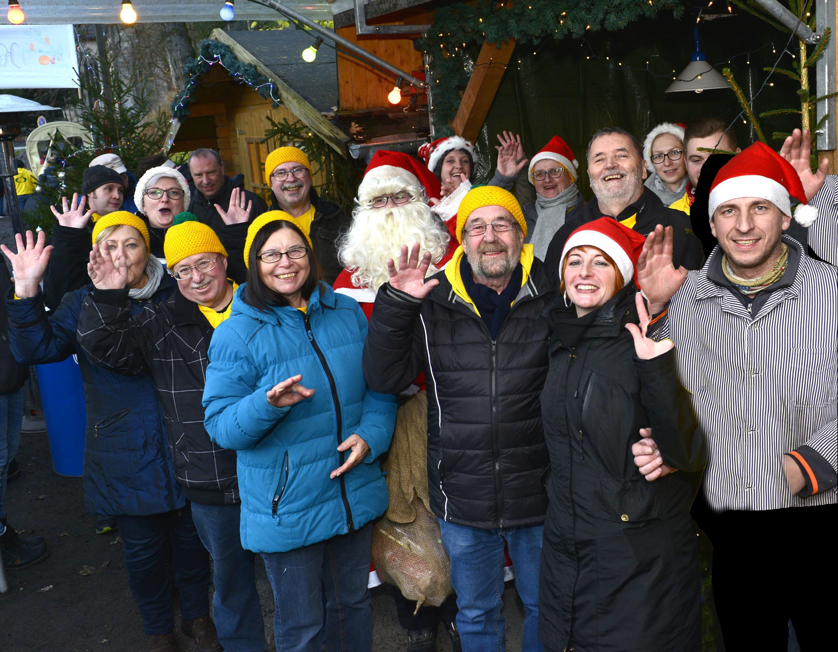 Viele Menschen winken auf einem Weihnachtsmarkt. In der Mitte steht der Weihnachtsmann.